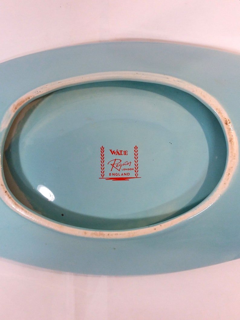 May include: A light blue oval ceramic dish with a white rim. The bottom of the dish has a red logo that reads "Wade Regalo London England" with a wreath on either side of the text.