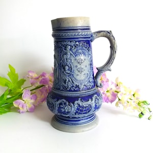 Puede incluir: Una jarra de cerámica azul y blanca con intrincados diseños en relieve, incluyendo un escudo central con un símbolo de átomo estilizado. La jarra tiene un asa y una base acampanada.