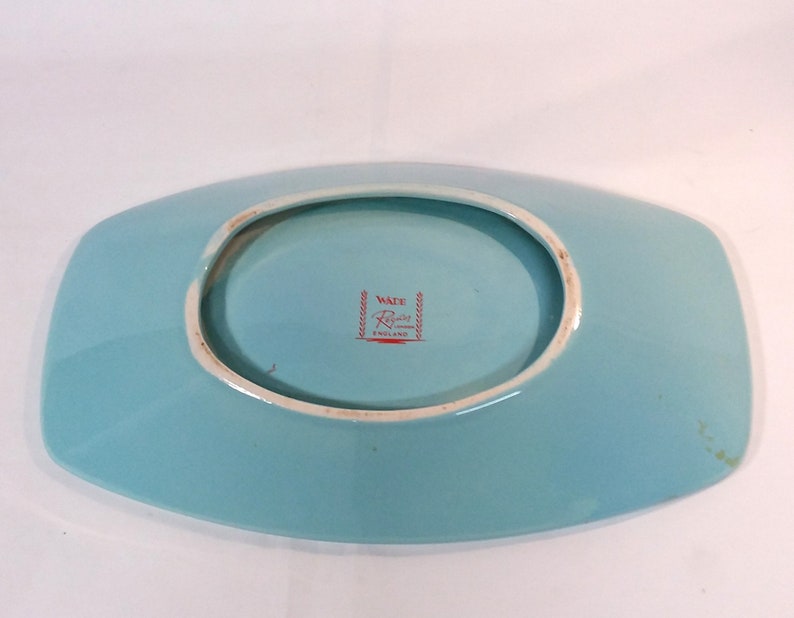May include: A light blue ceramic serving dish with a raised rim and a slightly oval shape. The bottom of the dish has a red and white label that reads "Wade Regent England".