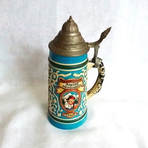 Könnte beinhalten: Ein blauer und weißer Bierkrug aus Keramik mit einem Metalldeckel und -griff. Der Krug zeigt eine farbenfrohe Illustration eines Mannes in traditioneller bayerischer Kleidung, der ein Bierglas hält. Der Text "Alpine Aygingerbrau Lager" ist auf den Krug gedruckt.
