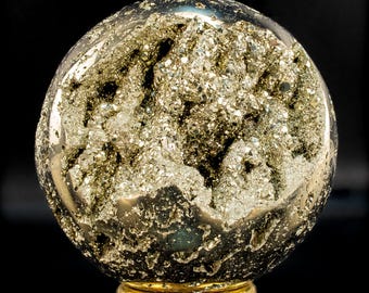 Pode incluir: Uma geodo de pirite esférica, com um exterior polido e um interior cristalino brilhante. A geodo repousa sobre uma base circular dourada, sobre um fundo preto. Os cristais de pirite brilham com brilho metálico.