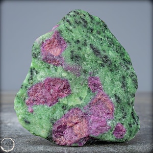 Op de afbeelding: Een ruwe, groene en roze edelsteen met een gemarmerd uiterlijk. De groene steen heeft donkergroene en zwarte vlekken. De roze gebieden zijn geconcentreerd in vlekken en hebben een donkerdere, roodachtige tint.
