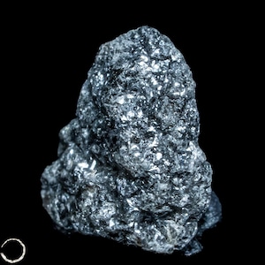 May include: A raw, dark gray mineral specimen with a highly textured, crystalline surface. The stone features numerous small, reflective facets that catch the light, creating a metallic, shimmering appearance against a solid black background.