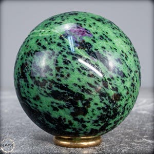 May include: A polished sphere of zoisite with ruby, displaying a vibrant green hue speckled with black and a touch of purple. The gemstone sits on a small, gold-colored base, showcasing its natural beauty.