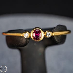 Könnte beinhalten: Ein goldener Ring mit einem ovalen Rubin-Mittelstein, flankiert von zwei kleineren, runden, klaren Edelsteinen. Der Ring wird auf einem schwarzen Ständer vor dunklem Hintergrund präsentiert. Das Ringband ist dünn und goldfarben.