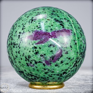 Può includere: Una sfera di pietra preziosa lucidata, prevalentemente verde con macchie nere e macchie di viola intenso. La sfera poggia su una base dorata, mettendo in risalto i motivi e i colori naturali della pietra. L'estetica generale è di bellezza naturale ed eleganza.