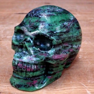 May include: A carved skull sculpture made of green, black, and pink stone. The skull has detailed features, including eye sockets, a nose cavity, and a full set of teeth. The stone has a marbled appearance, with streaks of colour throughout.