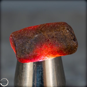 Könnte beinhalten: Ein grob geschliffener, rechteckiger Edelstein mit einer dunkelbraunen Außenseite und einem leuchtend roten Inneren, das von unten beleuchtet wird. Der Stein ist auf einem silbernen Metallständer montiert, der seine natürliche Textur und Farbvariationen zeigt. Die Kanten des Edelsteins sind abgerundet.