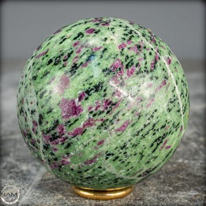 May include: A polished sphere of green zoisite with ruby inclusions, resting on a gold-colored base. The gemstone features a speckled pattern of deep pink and black throughout the green matrix. The sphere is a decorative item.