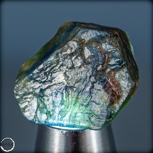 May include: A rough, faceted, blue-green gemstone with silver and brown inclusions. The gemstone is on a silver stand.