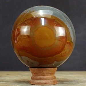 Op de afbeelding: Een gepolijste, veelkleurige agaatbol op een houten standaard. De bol heeft een groot, cirkelvormig, oranje-bruin patroon in het midden, omgeven door grijze en bruine banden.