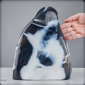 Peut inclure: Une sculpture en agate polie de forme libre, présentant un motif gris foncé et blanc. La pierre a une surface lisse et brillante et un sommet arrondi. Les zones blanches ressemblent à des nuages. La sculpture est exposée sur une surface claire.