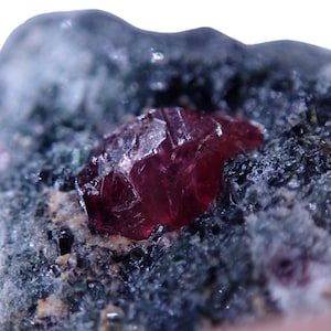 May include: A deep red ruby crystal with a faceted surface, embedded in a dark grey matrix.