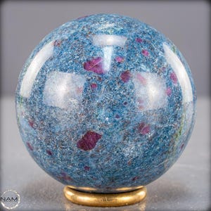 Peut inclure: Une sphère de pierre précieuse polie, principalement bleue avec des inclusions rouge rubis. La sphère repose sur une base circulaire dorée. La surface polie reflète la lumière, mettant en valeur les motifs et couleurs naturels. Diamètre d'environ 8 cm.