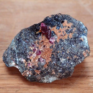 May include: A close-up of a dark grey rock with a cluster of bright red crystals. The crystals are embedded in the rock and have a sharp, pointed shape.