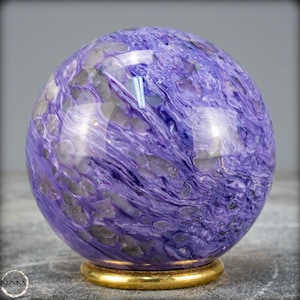 Pode incluir: Uma esfera polida de charoite, uma pedra ornamental. Apresenta padrões em espiral de cores roxas e cinzentas. A esfera repousa sobre uma pequena base dourada, realçando as suas cores naturais.