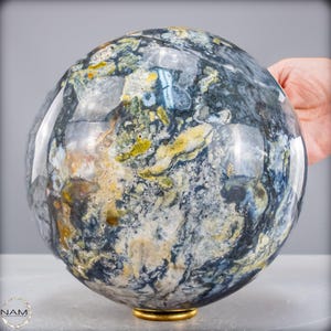 Puede incluir: Una gran esfera pulida con un patrón arremolinado de azul, gris, amarillo y blanco. La piedra preciosa se asienta sobre una pequeña base dorada. La esfera es sostenida por una mano, mostrando su tamaño y superficie reflectante.