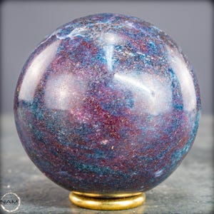 Peut inclure: Une sphère polie aux teintes bleues et violettes, posée sur un socle doré. La pierre présente une finition brillante, reflétant la lumière et mettant en valeur ses variations de couleurs uniques. Objet décoratif.
