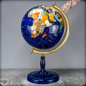Op de afbeelding: Een decoratieve wereldbol met een diepblauw oceaanoppervlak en ingelegde veelkleurige stenen continenten. De wereldbol is gemonteerd op een goudkleurige metalen meridiaanring en een donkerblauwe voet, voorzien van gedetailleerde geografische labels.
