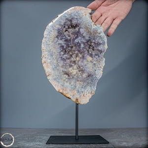 Könnte beinhalten: Eine große, runde, helllila und weiße Achat-Geoden-Scheibe auf einem schwarzen Metallständer. Die Geode hat eine natürliche, organische Form und eine glatte, polierte Oberfläche.