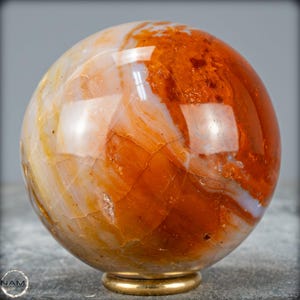 Op de afbeelding: Een gepolijste, bolvormige edelsteen met een mix van oranje, bruine en crèmekleurige tinten. De bol heeft een glanzende afwerking en staat op een kleine, goudkleurige basis. Het oppervlak van de steen vertoont natuurlijke patronen.