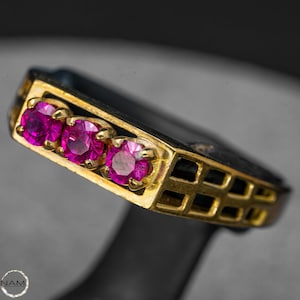Puede incluir: Primer plano de un anillo de oro con tres piedras preciosas rosas brillantes de talla ovalada. El anillo tiene una banda negra y un diseño de filigrana dorada en los laterales. Las piedras están engastadas en garras de oro.