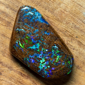 Könnte beinhalten: Ein brauner, grüner und blauer Opal-Edelstein mit einer rauen, natürlichen Form. Der Opal hat ein einzigartiges Muster aus wirbelnden Farben und eine dunkle, erdige Basis.