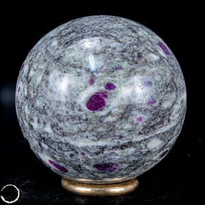 May include: A polished stone sphere with a gray and white marbled pattern, speckled with vibrant, deep pink ruby inclusions. The sphere rests on a small, gold-colored metal base, set against a black background.