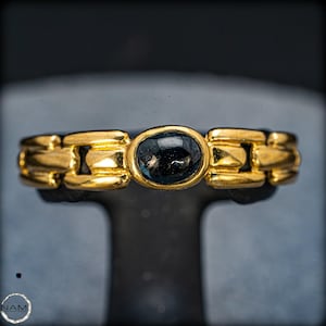 Puede incluir: Un anillo dorado con una piedra preciosa ovalada azul oscuro en el centro. El anillo presenta un diseño de eslabones de cadena. El anillo se muestra sobre un soporte negro sobre un fondo oscuro.