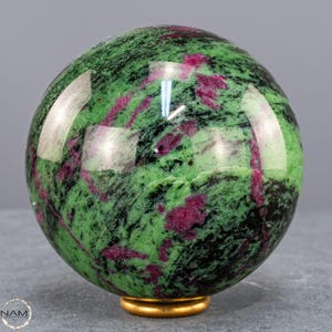Può includere: Una sfera lucidata di zoisite con inclusioni di rubino, che mostra una vibrante tonalità verde con striature di rosso scuro e nero. La gemma è posta su una piccola base dorata, che riflette la luce. Questa sfera decorativa è un oggetto d'arredo unico.