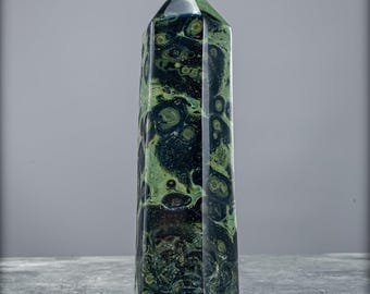 Puede incluir: Un obelisco de cristal pulido y alto con una parte superior puntiaguda. La piedra presenta un patrón moteado verde oscuro y negro. El obelisco está de pie sobre una superficie gris, con un fondo gris.
