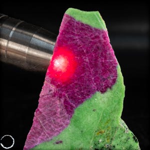 Pode incluir: Um close-up de uma pedra preciosa em bruto com uma paleta de cores vibrantes. A pedra apresenta um centro magenta profundo, rodeado por uma faixa verde, com uma luz vermelha brilhante a brilhar sobre a área magenta. A pedra preciosa está sobre um fundo preto.