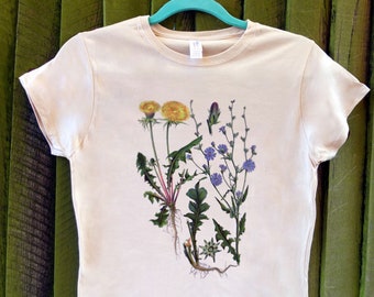 May include: A light beige t-shirt featuring a botanical illustration of wildflowers. The graphic includes yellow dandelions, purple flowers, green leaves, and visible roots. The shirt is hanging on a teal hanger against a green wooden background.