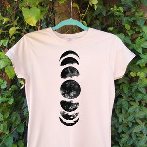 May include: A light beige t-shirt featuring a black watercolor design of the phases of the moon. The graphic shows the moon's cycle from crescent to full and back. The shirt is hanging on a light blue hanger against a backdrop of green foliage.