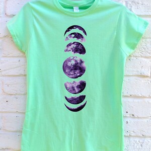 May include: A light green t-shirt featuring a purple moon phases design. The graphic shows the different stages of the moon, from crescent to full, in a vertical arrangement. The shirt is hanging on a teal hanger against a white brick wall.