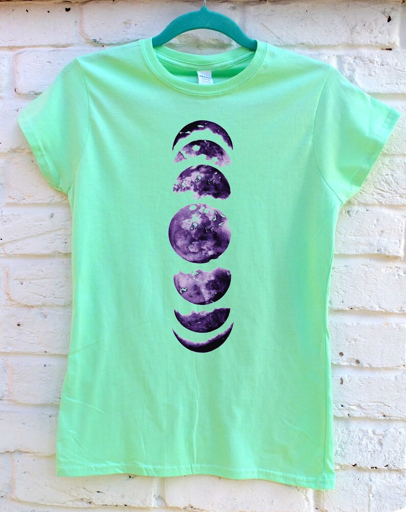 May include: A light green t-shirt featuring a purple moon phases design. The graphic shows the different stages of the moon, from crescent to full, in a vertical arrangement. The shirt is hanging on a teal hanger against a white brick wall.