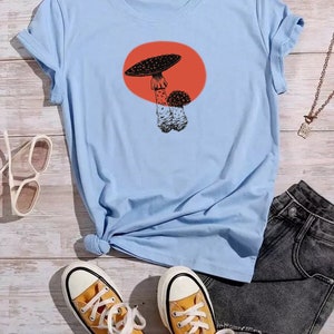 May include: Light blue t-shirt with a graphic of a mushroom in a red circle. The shirt is shown with a pair of yellow high-top sneakers and denim shorts.