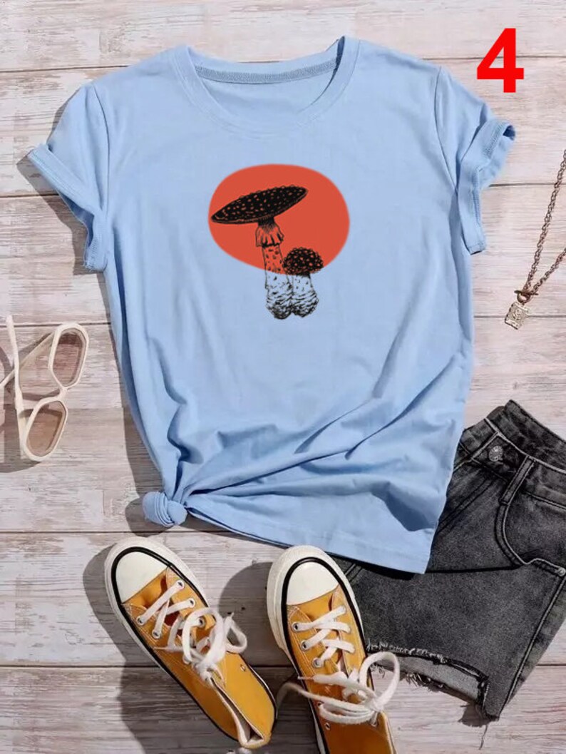 May include: Light blue t-shirt with a graphic of a mushroom in a red circle. The shirt is shown with a pair of yellow high-top sneakers and denim shorts.