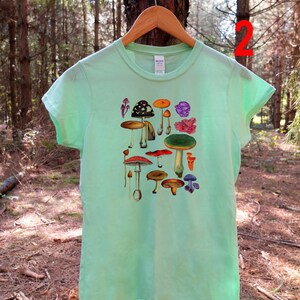 May include: A light green t-shirt with a colorful mushroom illustration. The shirt has short sleeves and a crew neck.