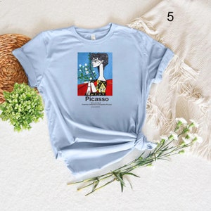 May include: Light blue t-shirt featuring a graphic print of a painting by Pablo Picasso. The painting depicts a woman wearing a red and yellow dress with white flowers. The text "Picasso" and "The Late Works" is printed below the painting.