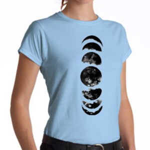 May include: Light blue t-shirt featuring a vertical black and white watercolor design of the phases of the moon. The design includes crescent, half, and full moon shapes. The shirt has short sleeves and a crew neck.