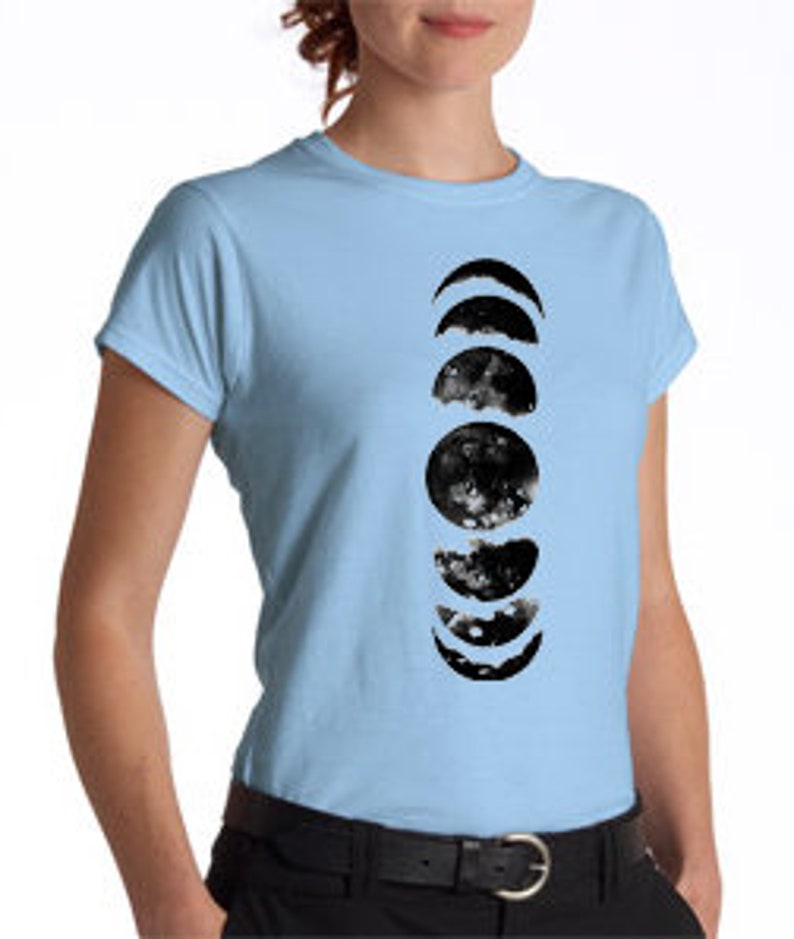 May include: Light blue t-shirt featuring a vertical black and white watercolor design of the phases of the moon. The design includes crescent, half, and full moon shapes. The shirt has short sleeves and a crew neck.