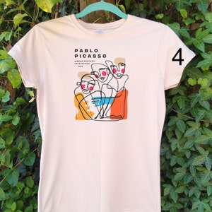 May include: A beige t-shirt with a graphic print of a line drawing of three faces. The text "PABLO PICASSO WOMAN PORTRAIT IMAGINAIRES 1962" is printed on the shirt. The number "4" is printed on the right sleeve.