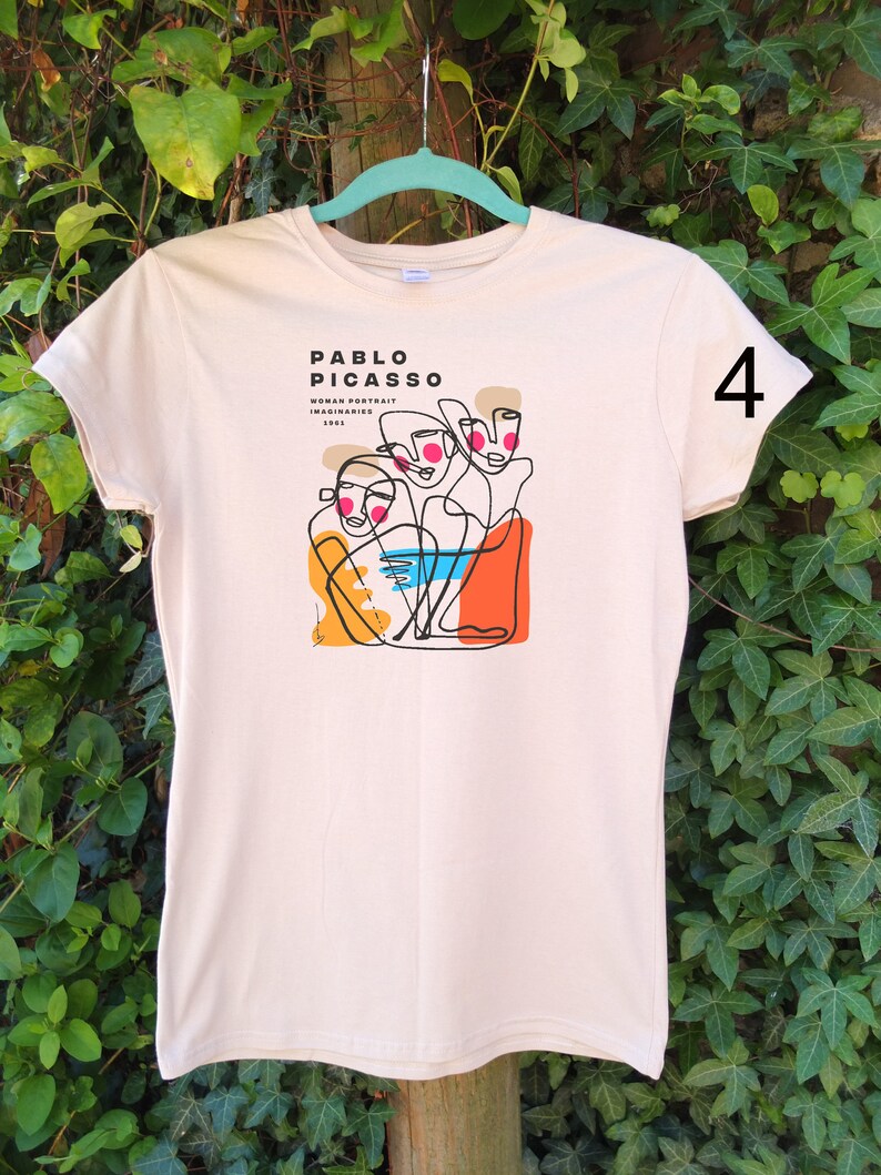May include: A beige t-shirt with a graphic print of a line drawing of three faces. The text "PABLO PICASSO WOMAN PORTRAIT IMAGINAIRES 1962" is printed on the shirt. The number "4" is printed on the right sleeve.