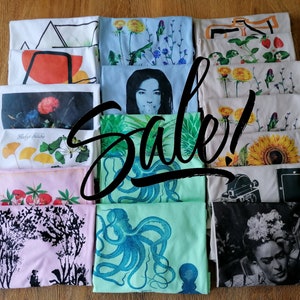 Puede incluir: Una pila de 10 camisetas diferentes con varios diseños, incluyendo patrones florales, un girasol, un pulpo y un retrato de Frida Kahlo. Las camisetas son todas blancas con diseños negros. La palabra "¡Venta!" está escrita en negro en el medio de la imagen.