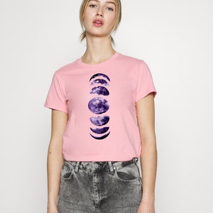 May include: A light pink t-shirt featuring a graphic of the phases of the moon in shades of purple and blue. The shirt is a crew neck style and is paired with gray acid wash jeans.