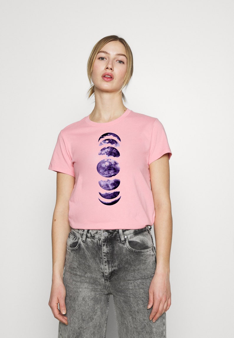 May include: A light pink t-shirt featuring a graphic of the phases of the moon in shades of purple and blue. The shirt is a crew neck style and is paired with gray acid wash jeans.
