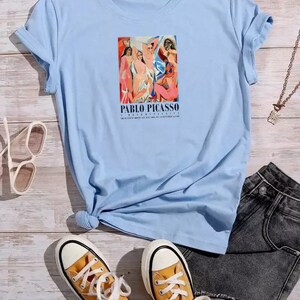 May include: Light blue t-shirt featuring a graphic print of a painting by Pablo Picasso. The painting depicts a group of people in a colourful abstract style. The text "PABLO PICASSO" is printed below the painting.