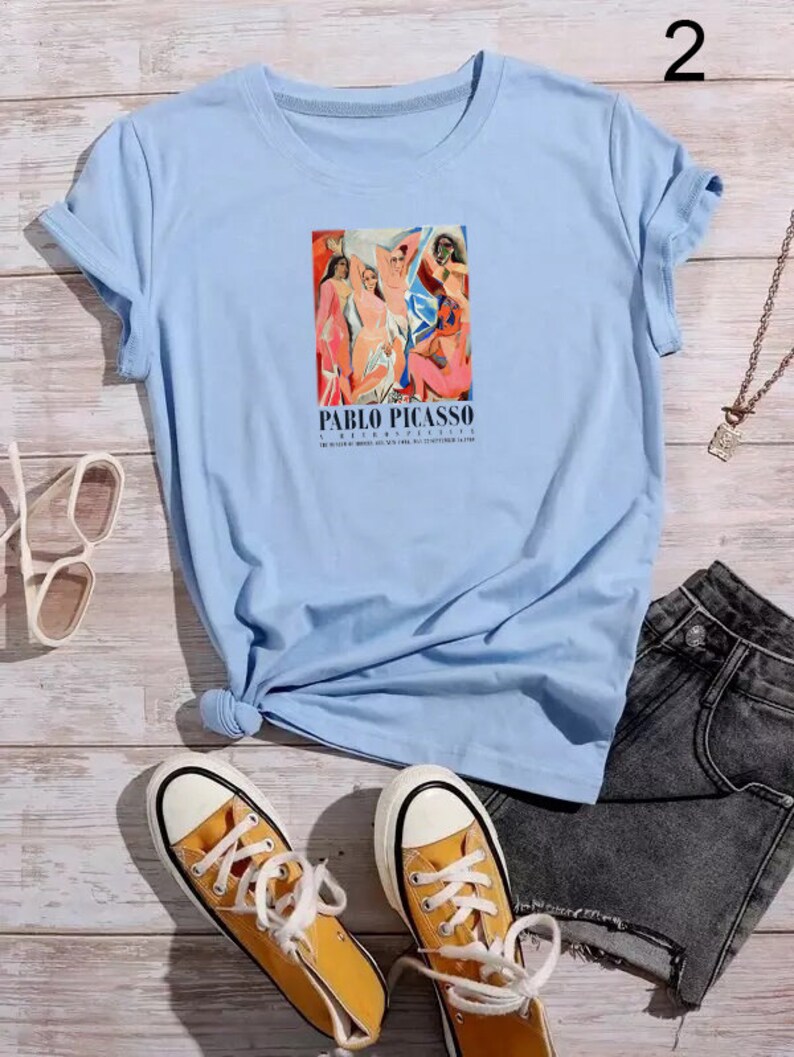 May include: Light blue t-shirt featuring a graphic print of a painting by Pablo Picasso. The painting depicts a group of people in a colourful abstract style. The text "PABLO PICASSO" is printed below the painting.