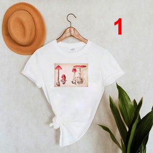 May include: White t-shirt with a vintage illustration of red mushrooms on the front. The illustration is in a rectangular frame.